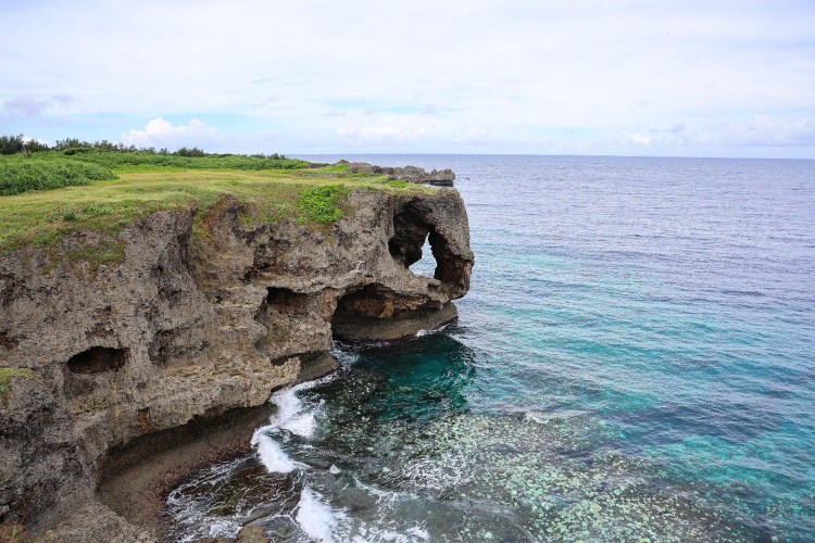 Scenic view of a rocky coastal cliff with a natural arch, overlooking calm turquoise waters and a clear sky.