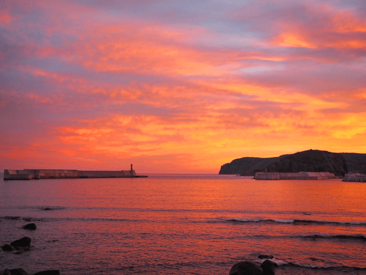Vibrant sunset over the ocean with silhouettes of breakwaters and a lighthouse in the foreground.